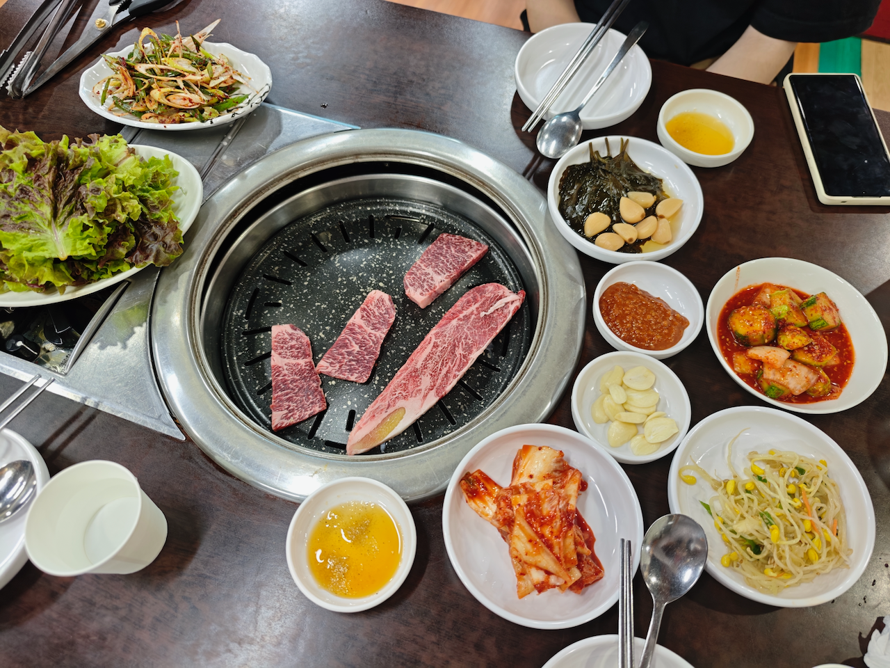 烤肉桌：生菜、蒜片、辣椒酱、泡菜、豆芽围着一圈，中间是还在冒烟的烤盘
